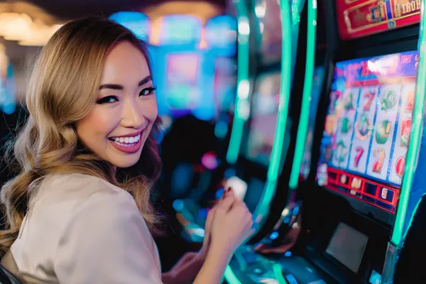 A woman smiling by bright slot machines showing lucky symbols, showcasing the exciting slot offerings at SPINARAA.